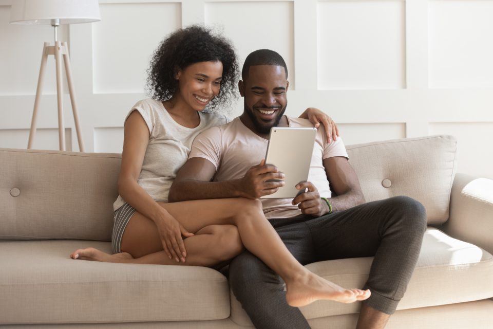 African couple in home clothes sit together on sofa embracing spend lazy weekend using gadget tablet, booking hotel planning vacation, watching videos enjoy free time, entertaining in internet concept
