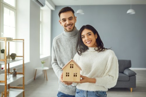 Happy couple holding a mock up of a house in their hands while standing in a room at home.