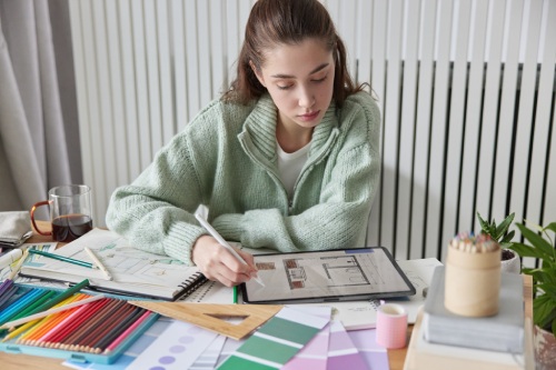 Indoor shot of young female interior designer works on apartment design project draws in digital tablet with stylus thinks about decorations of apartment wears casual jumper poses at desktop.