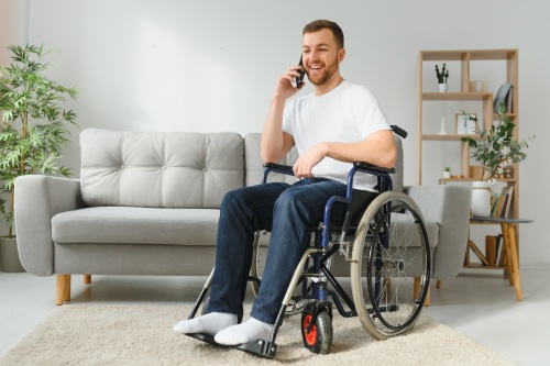 The disabled person sits in a wheelchair. He is talking to someone on his smartphone. He is in his large bright living room. He smiles.