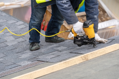 Real photo of professional roofer worker in uniform work wear using air or pneumatic nail gun and installing asphalt or bitumen shingle on top of the new roof under construction residential building