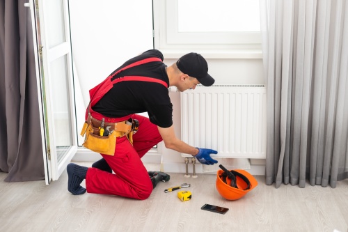 Man in work overalls using wrench while installing heating radiator in room. Young plumber installing heating system in apartment. Concept of radiator installation, plumbing works and home renovation