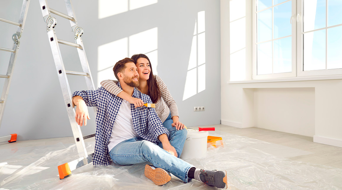 A couple perched on a ladder in a vacant room, discussing their plans for remodeling versus renovating the space.