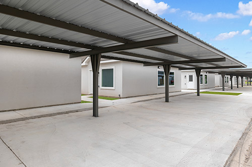 Covered walkway with a metal roof adjacent to a white building, featuring Tri-Plex, Four-Plex, and Six-Plex homes in Texas.