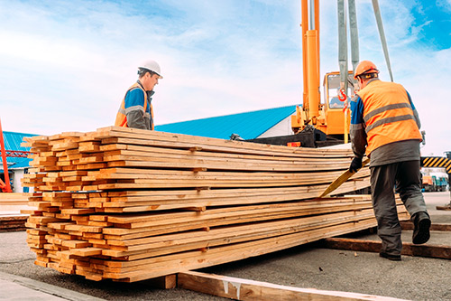 Two men wearing orange vests are actively sorting through a pile of wood in a work area.