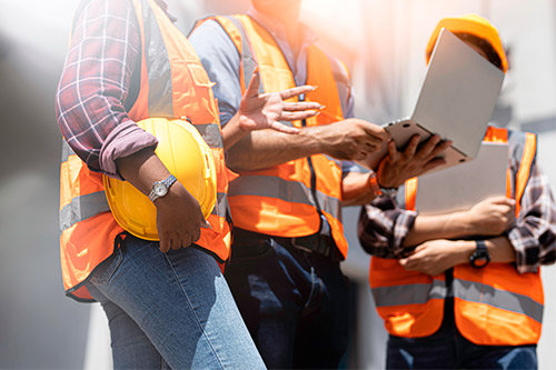 Three construction workers wearing orange vests and hard hats discuss plans, emphasizing the role of construction in business growth.
