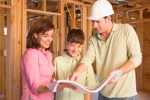 A family of three stands together on a construction site, wearing hard hats and safety vests